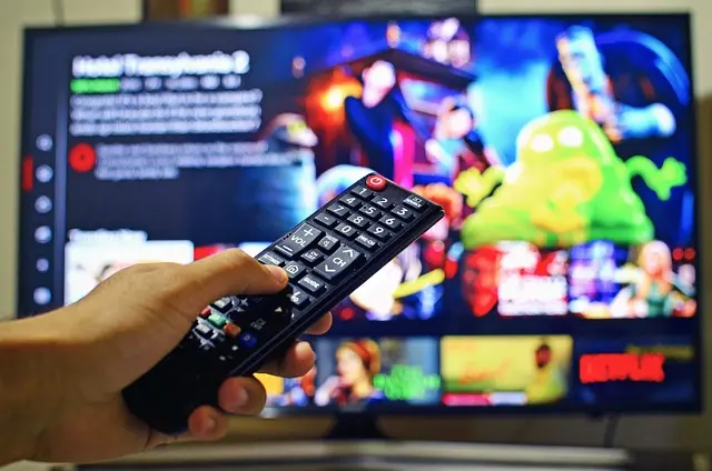Hand holding TV remote control pointing at colorful streaming interface displaying various entertainment content through IPTV subscriptions